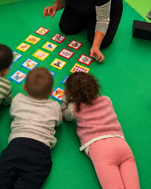 Tre bambini sdraiati sul tappeto della Casa giocano con le carte illustrate insieme all'insegnante, durante una lezione di Baby inglese