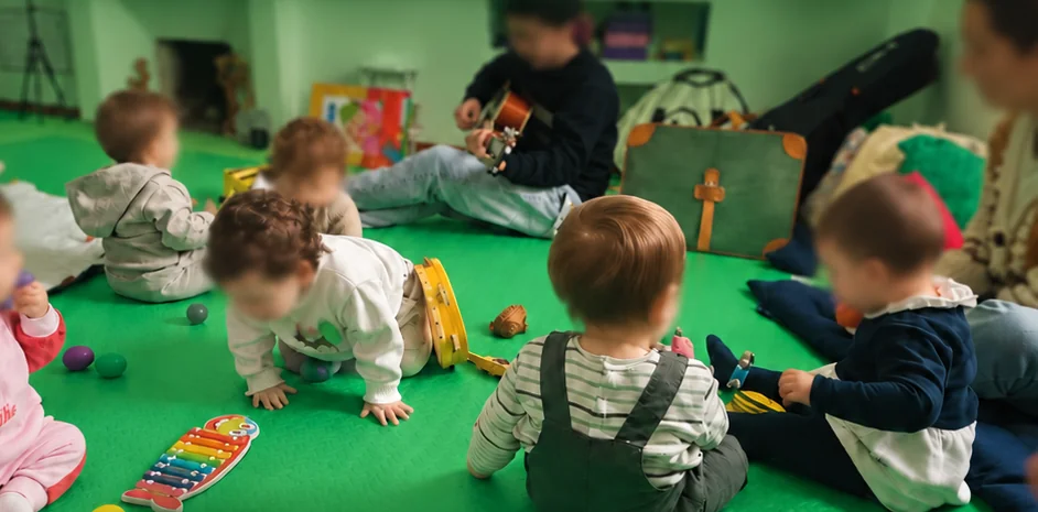 Bambini piccoli sul tappeto della Casa con sonagli, tamburelli e uno xilofono, mentre il maestro suona la chitarra, durante una lezione di Baby musica
