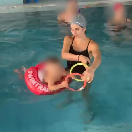 Un bambino nel salvagente gioca con gli anelli colorati insieme all'istruttrice, con altre mamme in acqua intorno