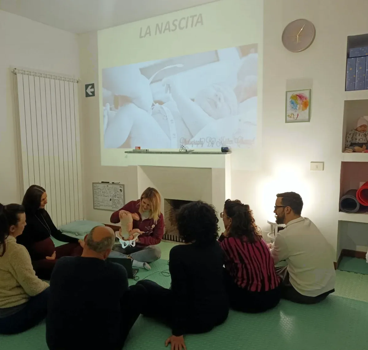 Un incontro del corso preparto in Casa, con le coppie sedute in cerchio e l'ostetrica che mostra come nasce un bambino